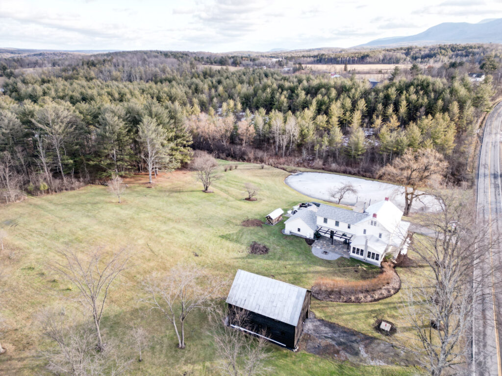 Aerial view of DeerWatch property showcasing the barn, home, and pond. Mountains are seen in the distance as well as a coniferous forest.